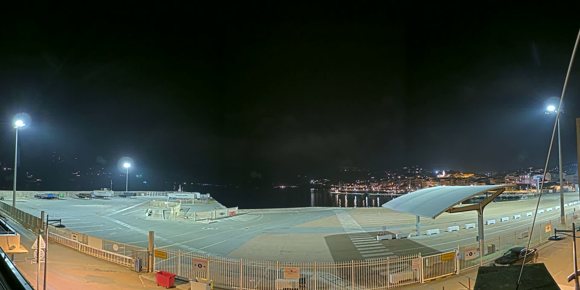 A nighttime view of an empty port area with a few parked vehicles, illuminated by bright lights, overlooks a calm body of water and a city skyline in the distance.