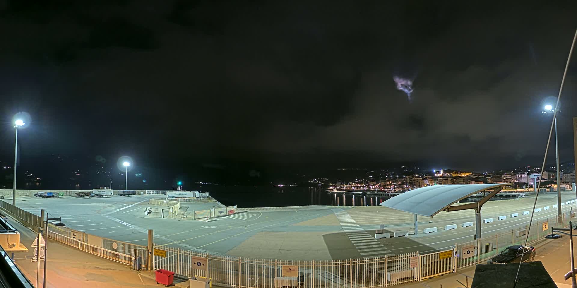 A nighttime view of an empty port area with several lights illuminating a large paved lot, overlooked by a town in the distance under a dark, cloudy sky.