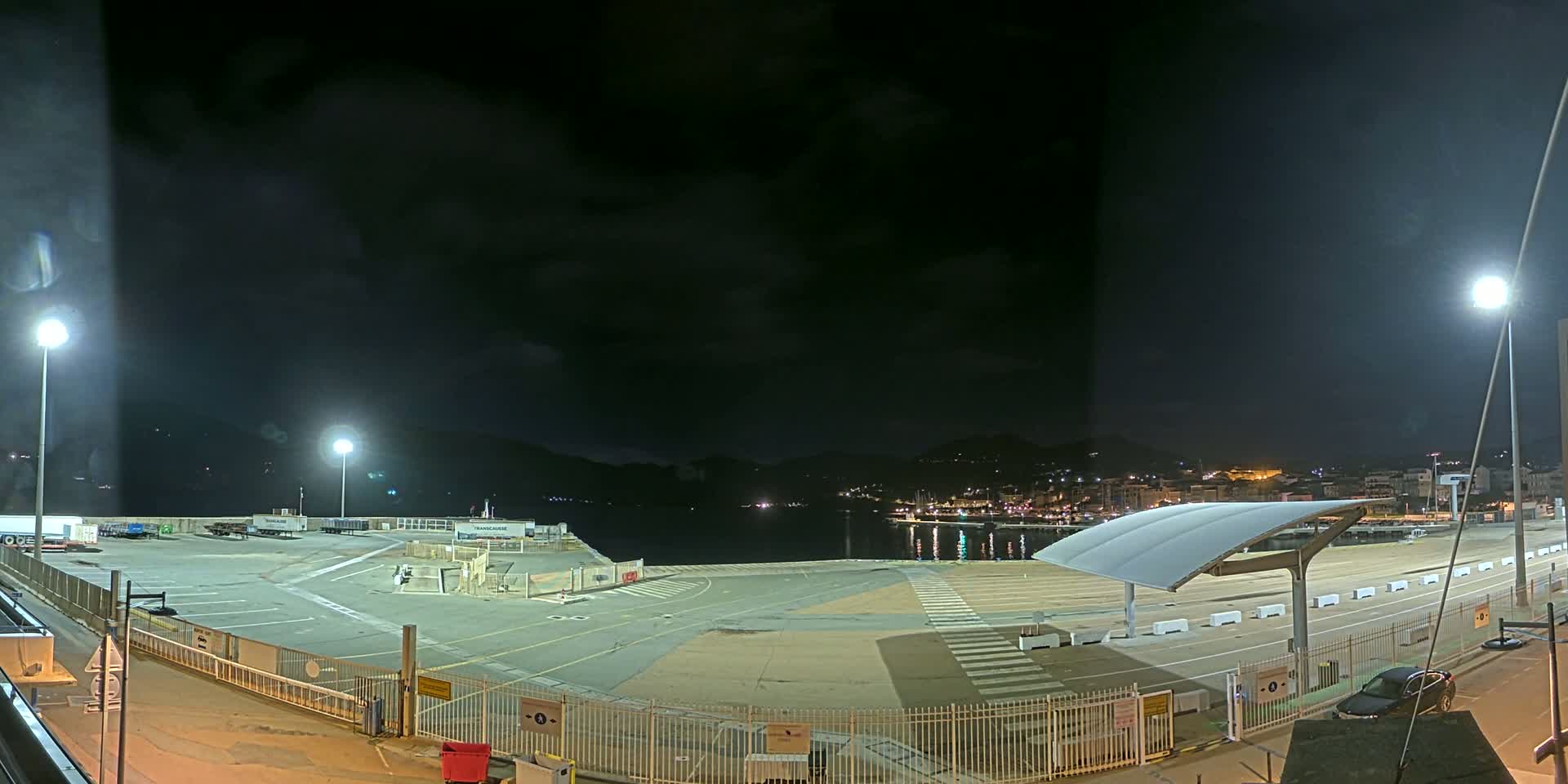 An empty, brightly lit port at night under a mostly cloudy sky, with a town visible across the water.