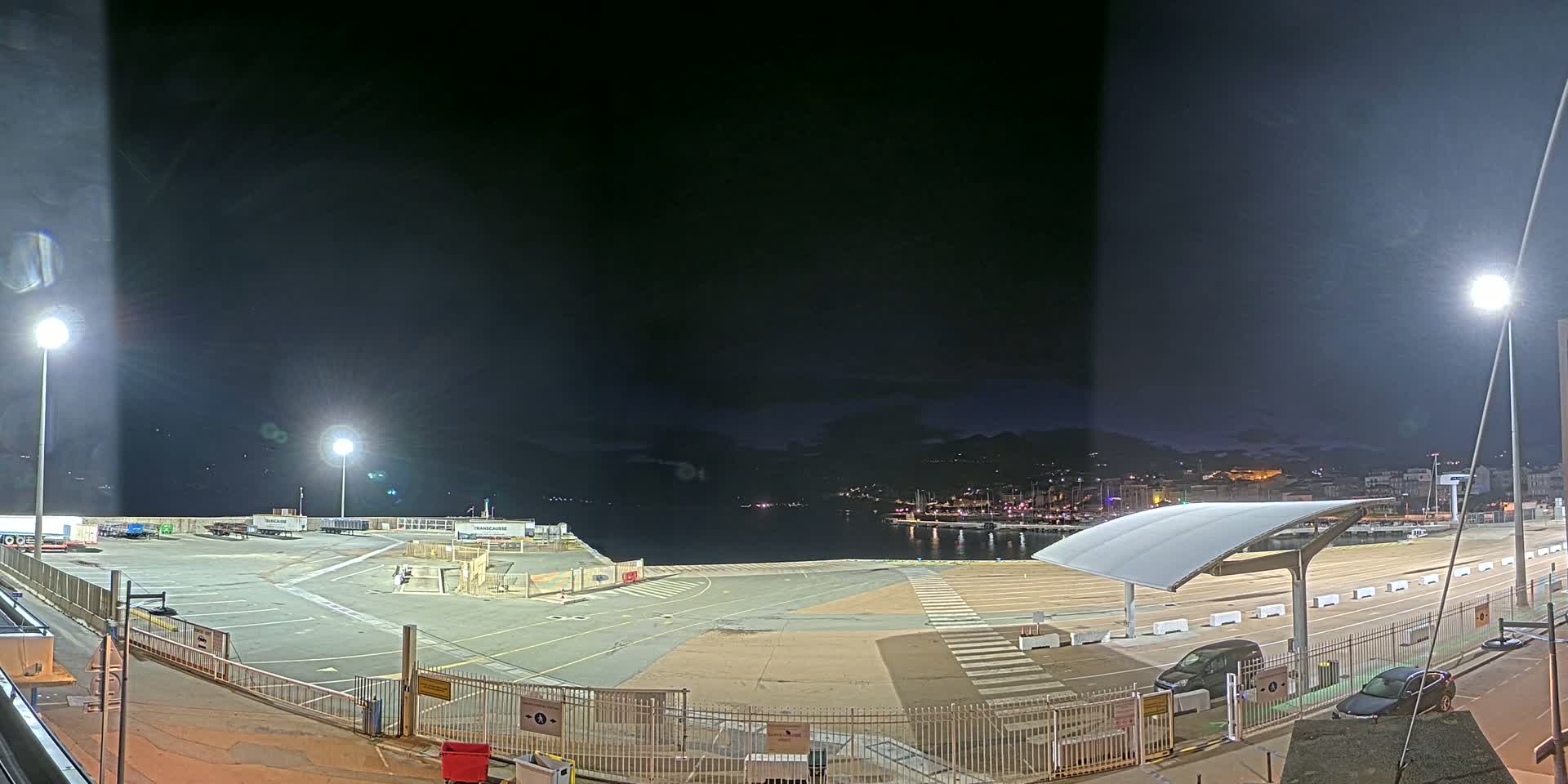 A brightly lit nighttime harbor area with a mostly empty paved lot, a few cars, and a distant city skyline visible across the water under a dark, clear sky.