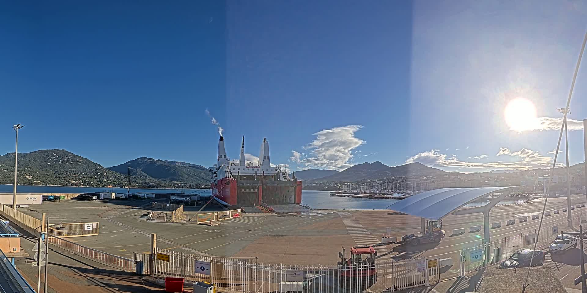 A large red and white ferry is docked at a port on a sunny day, with mountains in the background and several vehicles parked nearby.