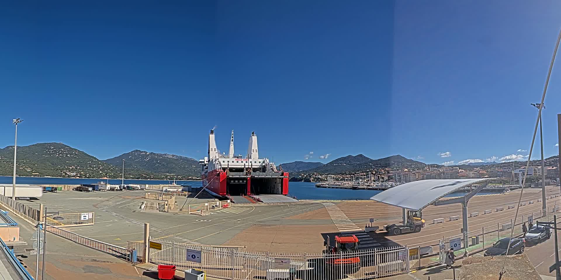 A large red and white ferry is docked at a port on a sunny day, with mountains visible in the background.