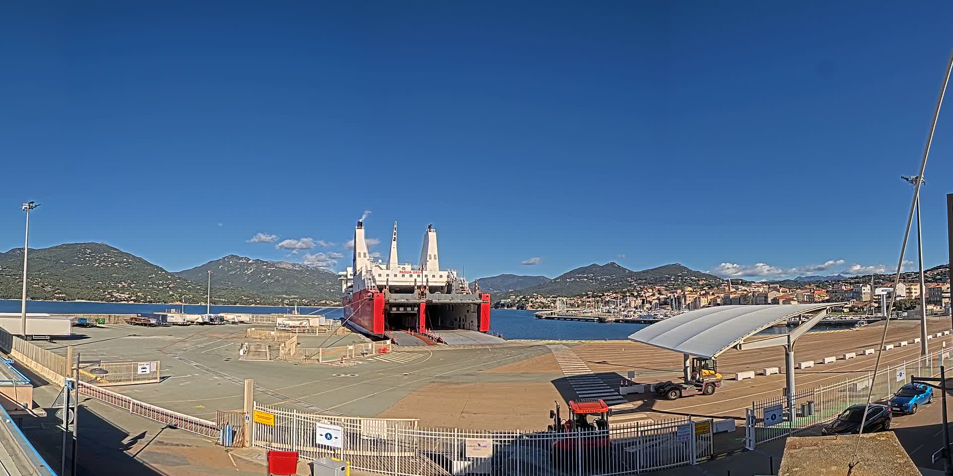 A large red and white cargo ship is docked at a port under a clear blue sky, with mountains in the background.