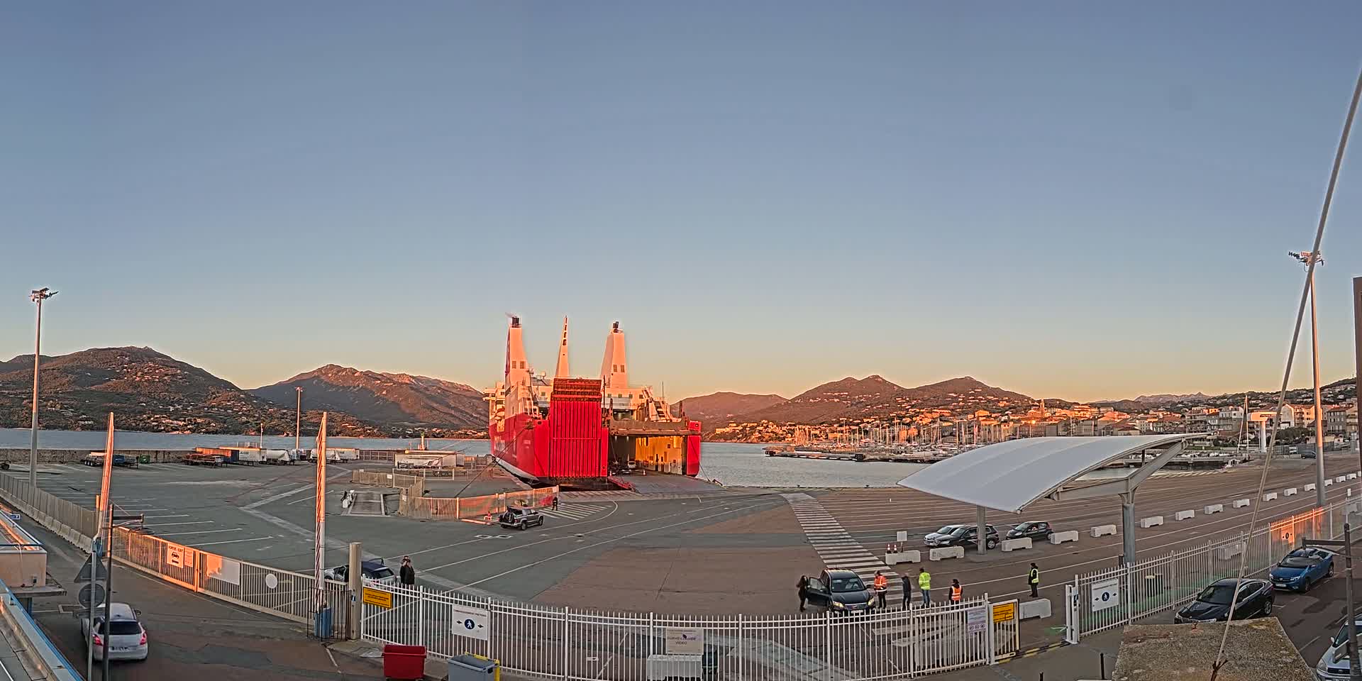 A large red ferry is docked at a port, with cars and people near it, under a clear sunset sky and mountains in the background.