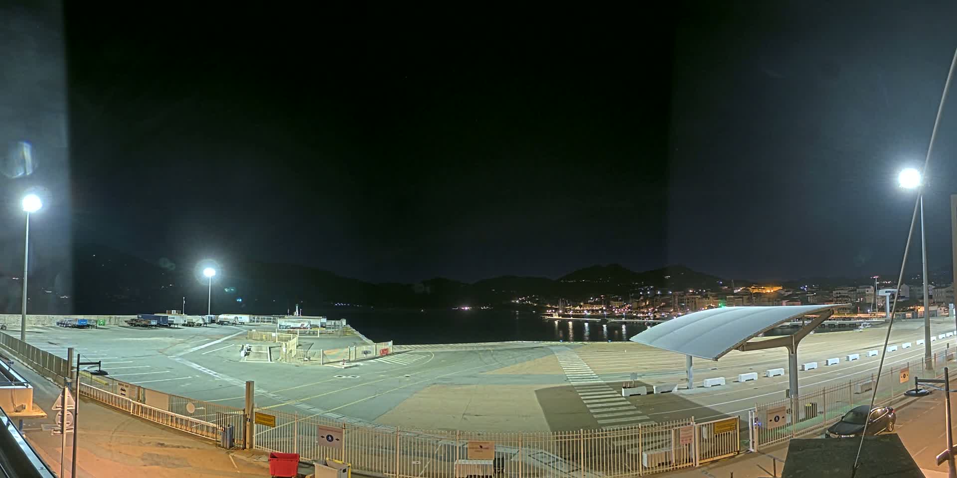 A nighttime view of an empty port area with a few parked vehicles, illuminated by several bright lights, overlooking a calm body of water and a distant town nestled against hills.