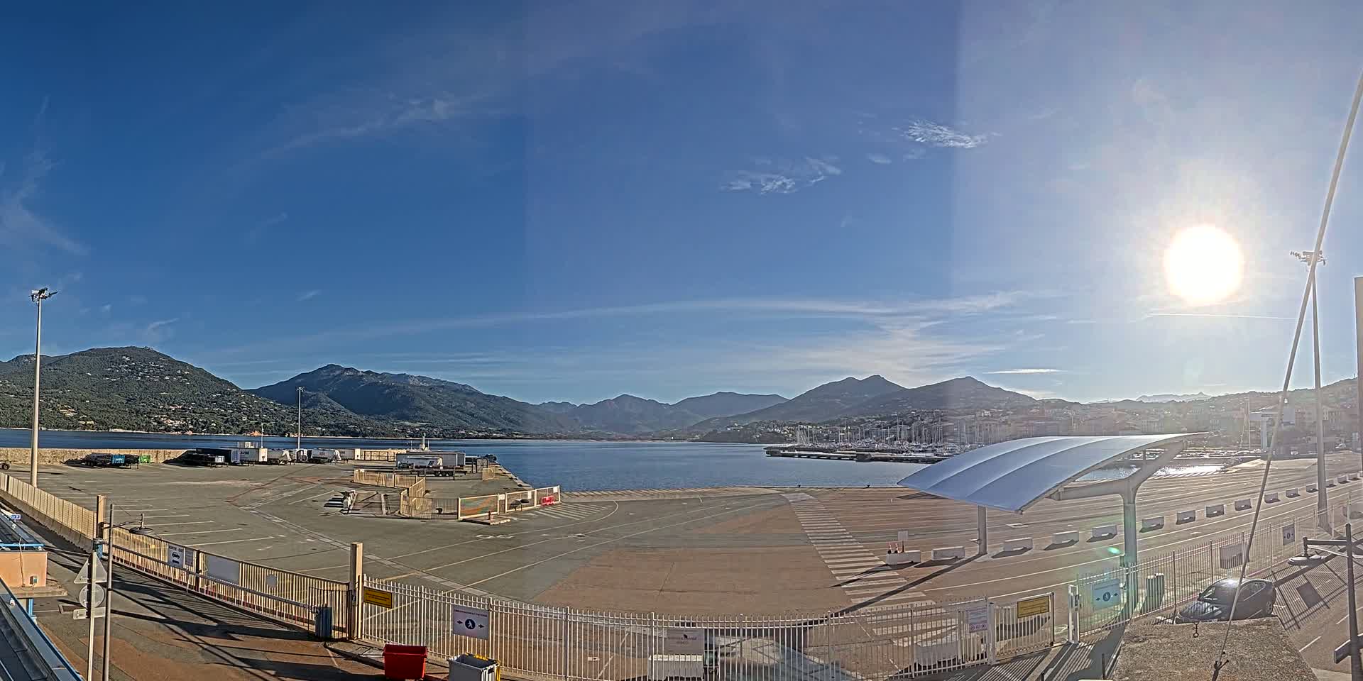 A sunny day at a port with a calm body of water, mountains in the background, and an empty parking lot in the foreground.