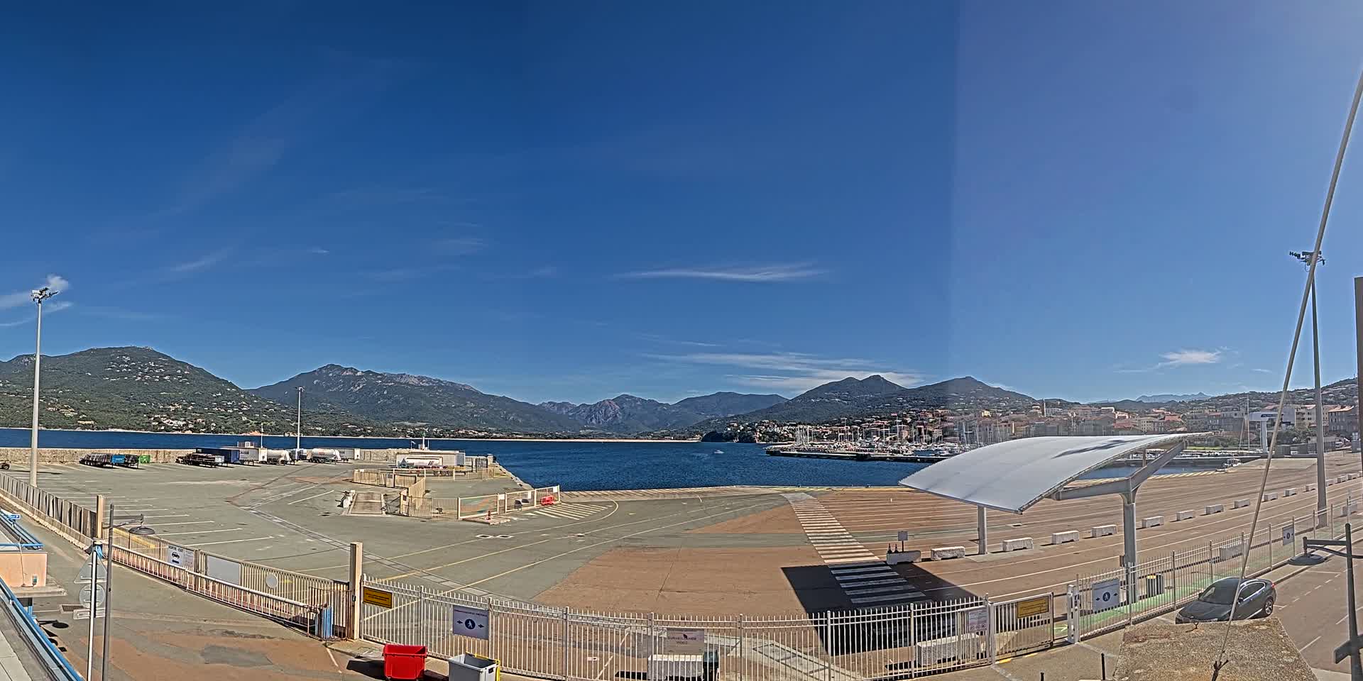 A mostly empty port area with a few vehicles, bordered by a metal fence, overlooks a calm body of water and mountains under a clear blue sky.