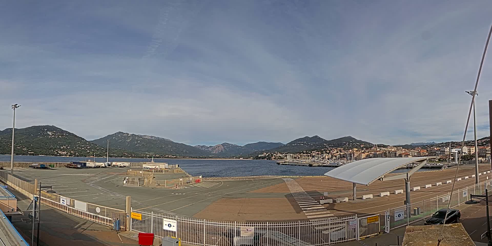 A mostly empty port area with a calm body of water and mountains in the background under a mostly clear sky.