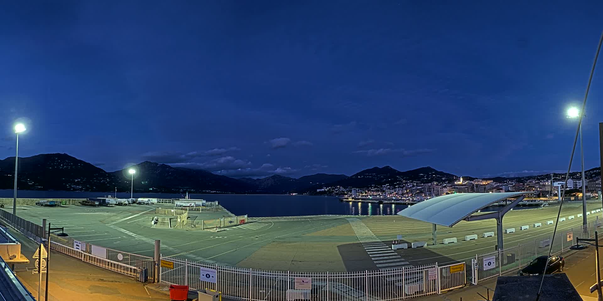 A mostly empty, illuminated port area at twilight, with mountains and a town visible across the water under a clear, dark blue sky.