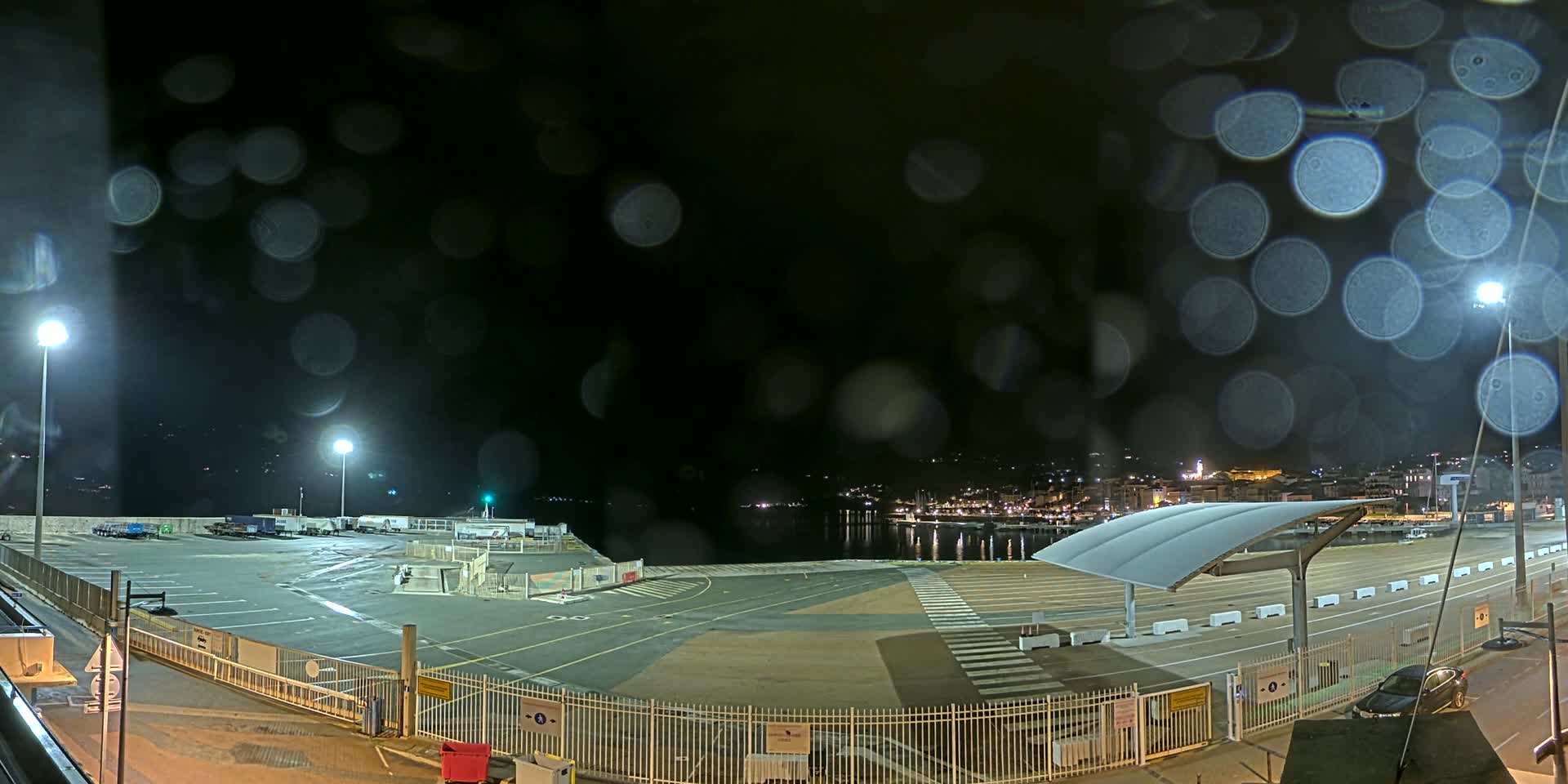 A nighttime view of an empty port area with a few vehicles, illuminated by lights, seen through rain-covered glass.