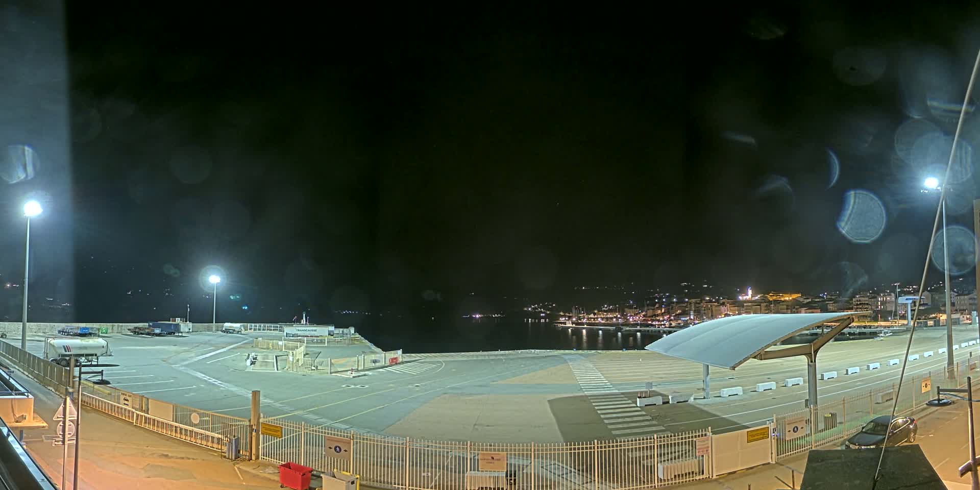 An empty, brightly lit nighttime port area with a few vehicles, overlooked by a city across the water, and viewed through rain-streaked glass.