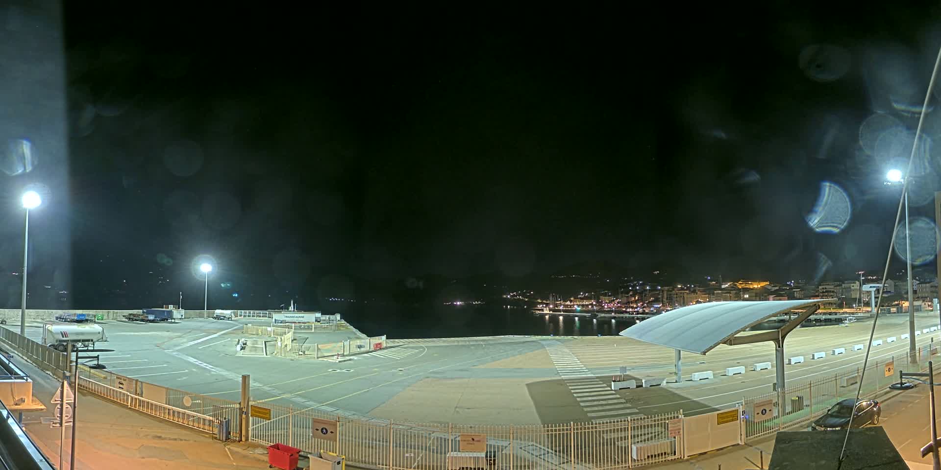 A nighttime view of an empty port area with a few parked vehicles, lit by several lamps, under a dark, slightly misty sky.