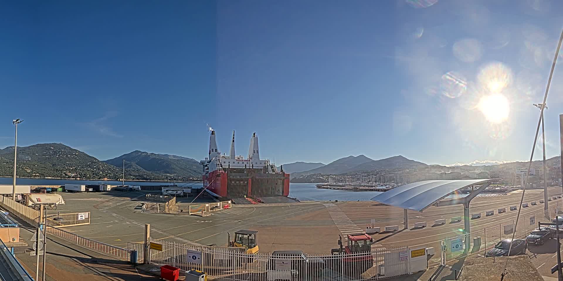 A large red and white cargo ship is docked at a port, under a bright sunny sky, with mountains in the background.