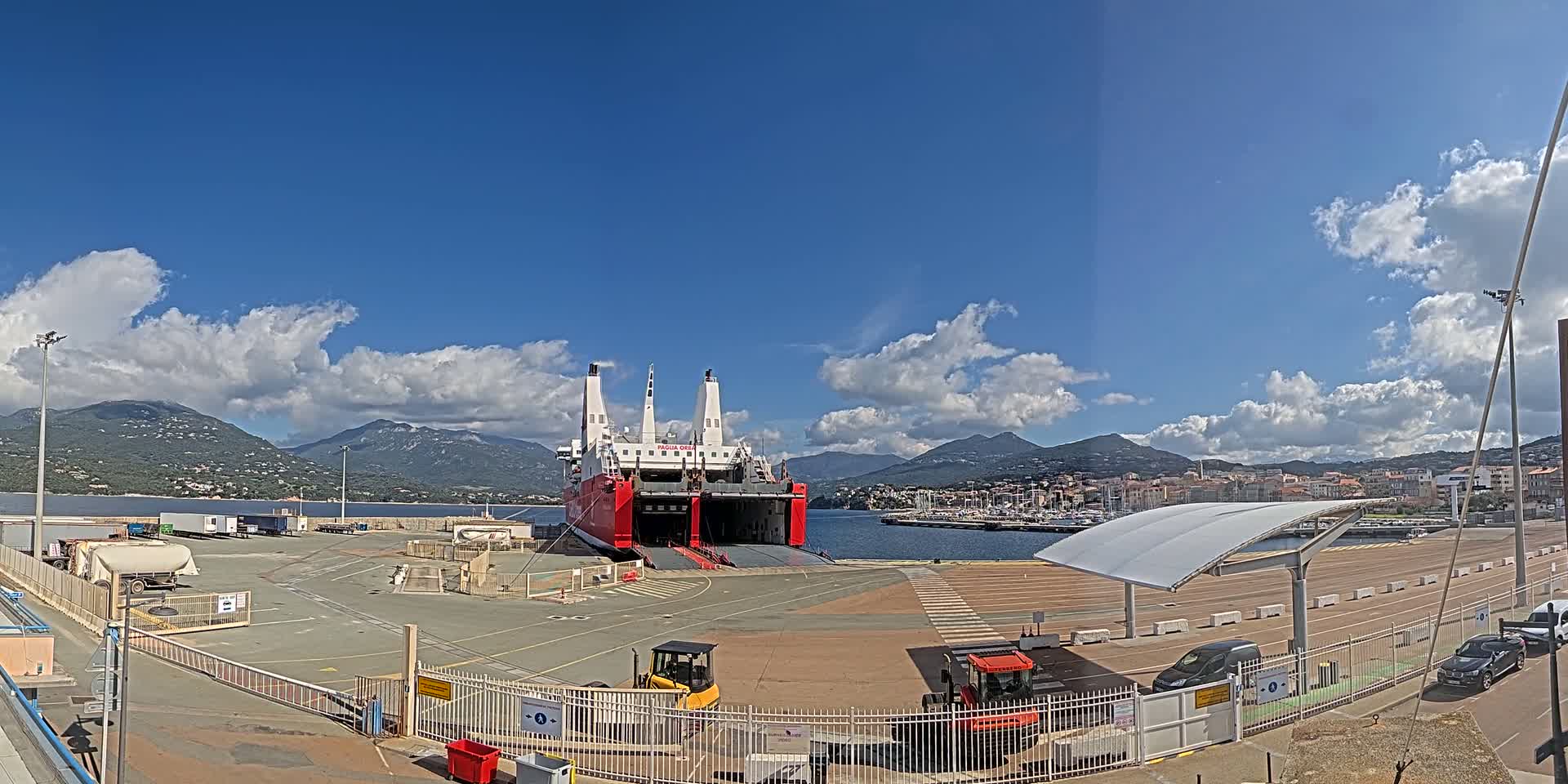 A large red and white ferry is docked at a port under a mostly sunny sky with scattered clouds, mountains in the background.