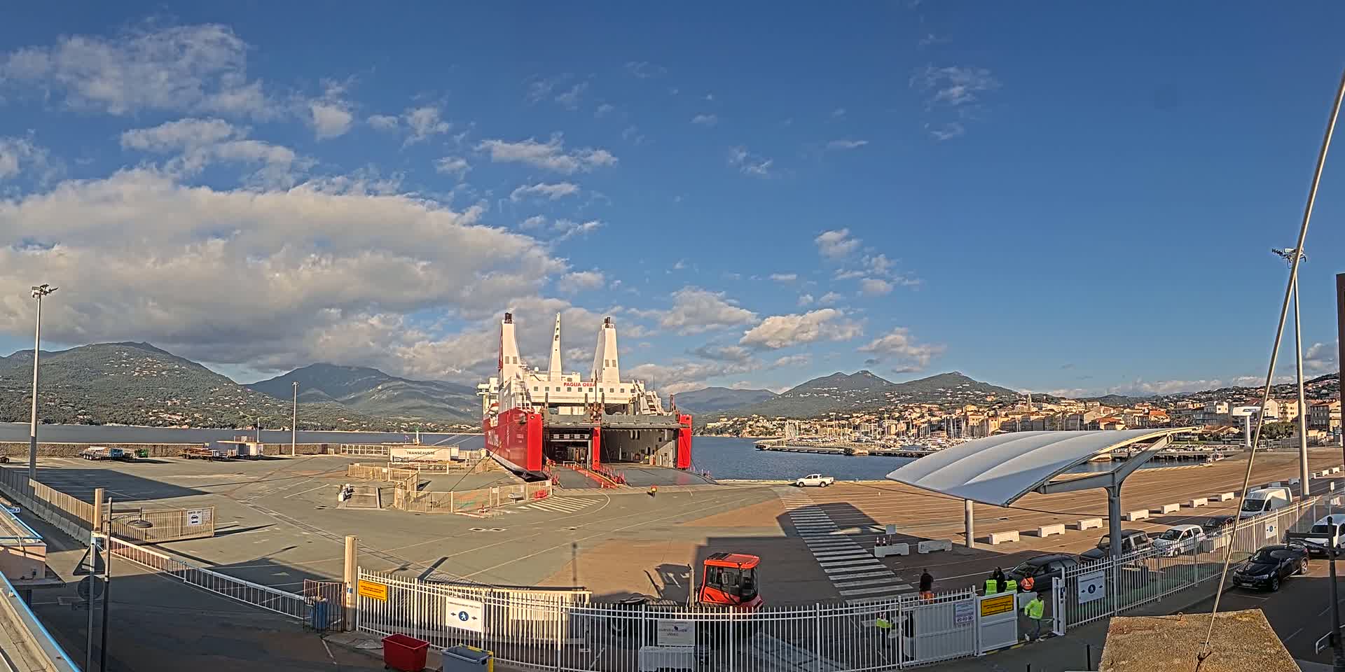 A large red and white ferry is docked at a port under a mostly sunny sky, with mountains and a town visible in the background.