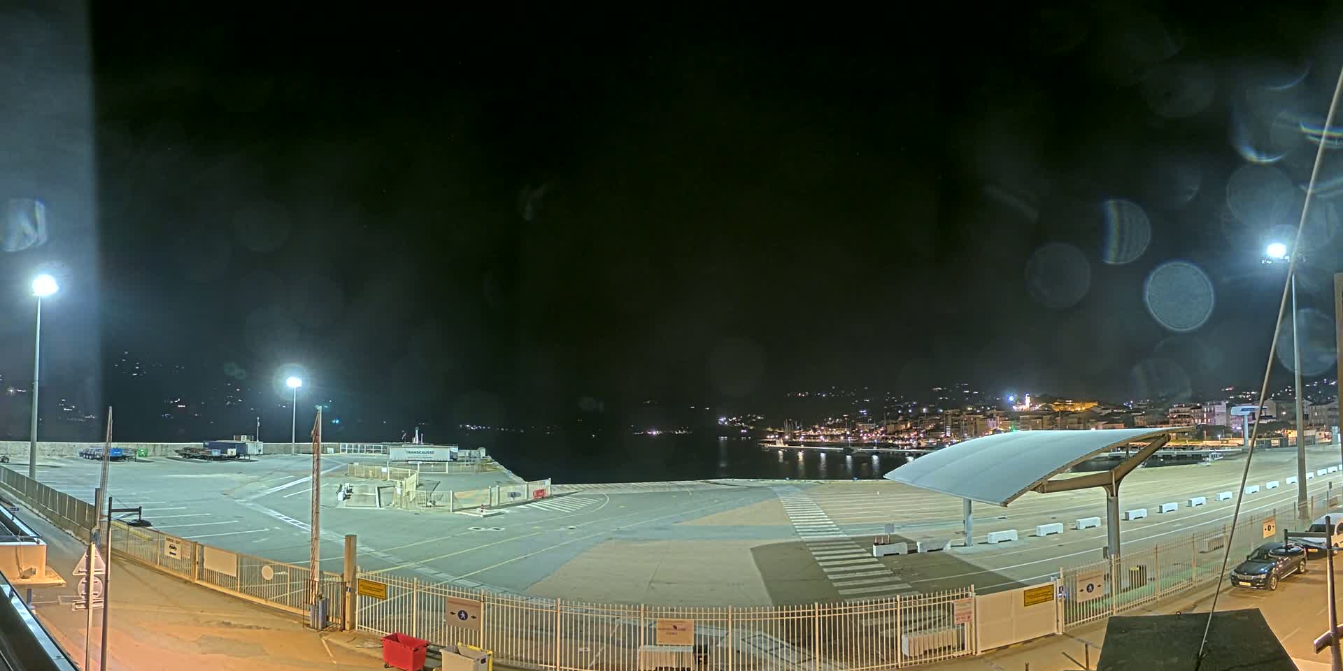 A nighttime view of an empty port area with a few vehicles, illuminated by several lights, overlooking a calm body of water with a town visible in the distance under a dark, slightly hazy sky.