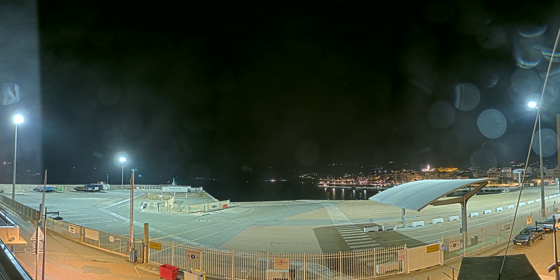 An empty, illuminated port area at night with rain on the camera lens, overlooking a calm body of water and a town in the distance.