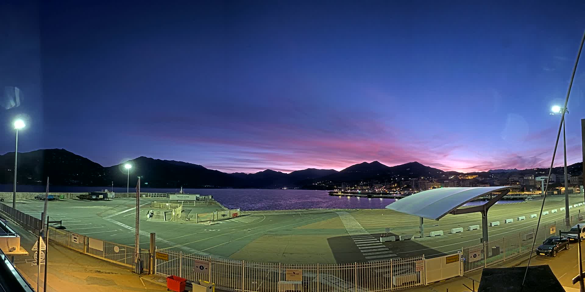 An empty port area with a modern canopy sits under a twilight sky, mountains in the background exhibiting a purplish hue at sunset.