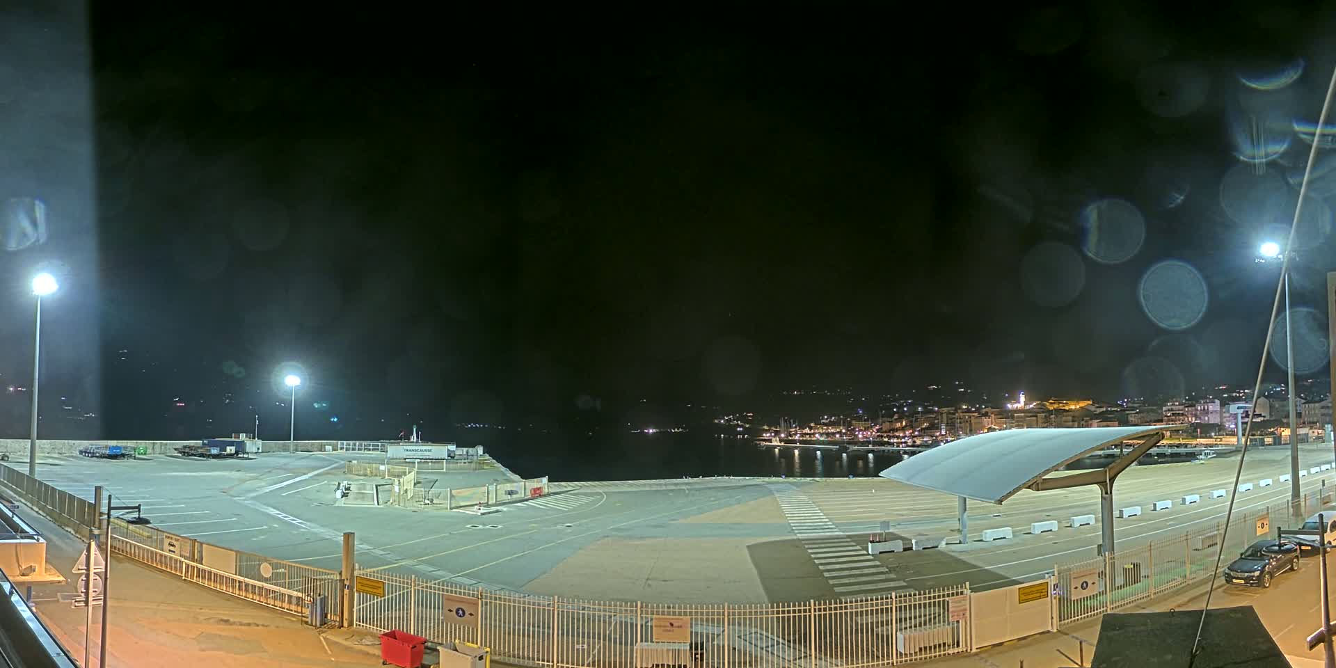 An empty, brightly lit port area at night, viewed from above, with a town visible across the water, and rain or condensation on the camera lens.