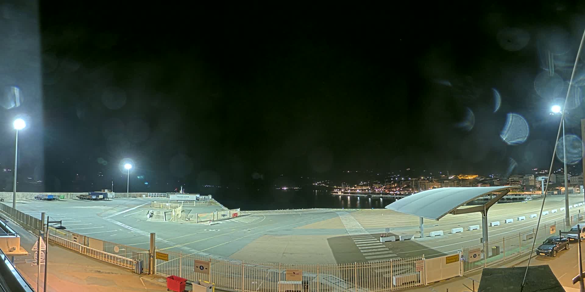 A nighttime view of an empty port area with a few vehicles parked, illuminated by several bright lights, under a dark, slightly rainy sky.