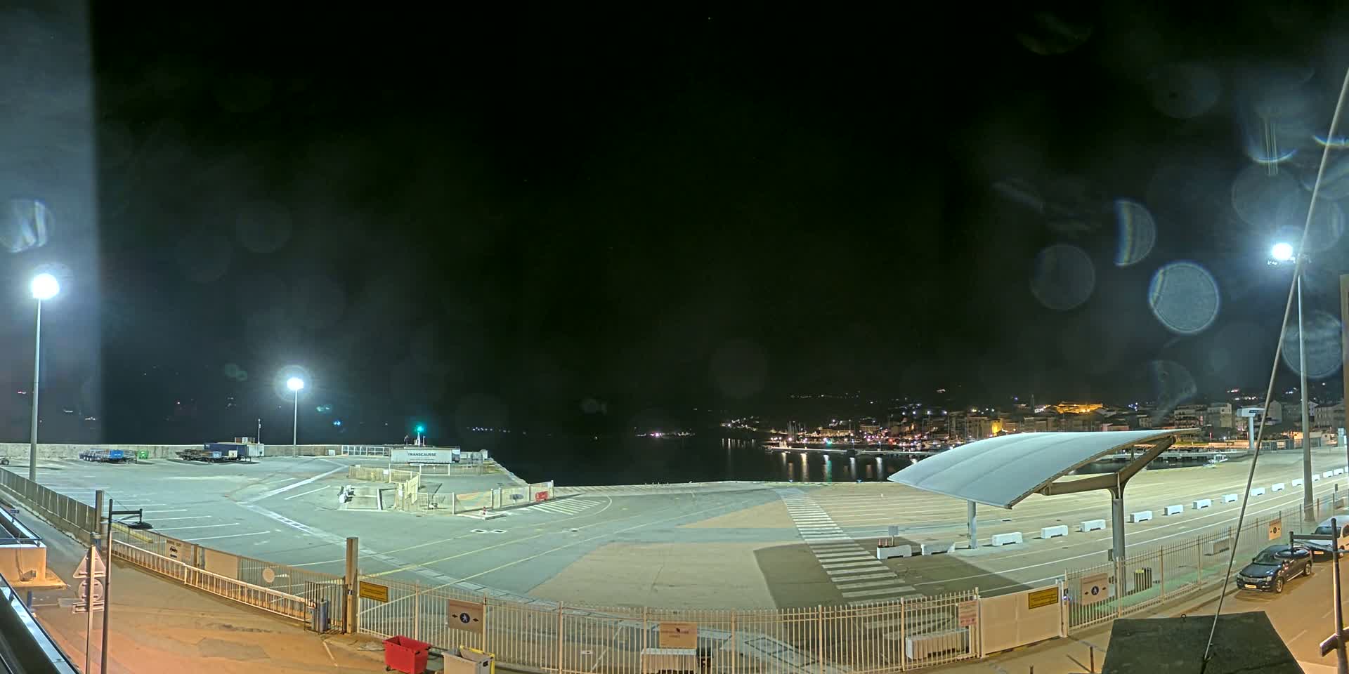 An empty, nighttime port area with a covered waiting area, a few parked vehicles, and a town visible across the water under a dark, possibly rainy sky.
