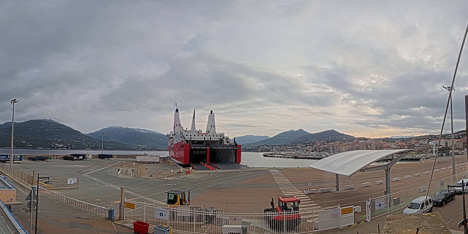 A large red and white ferry is docked at a port, with overcast skies and mountains in the background.
