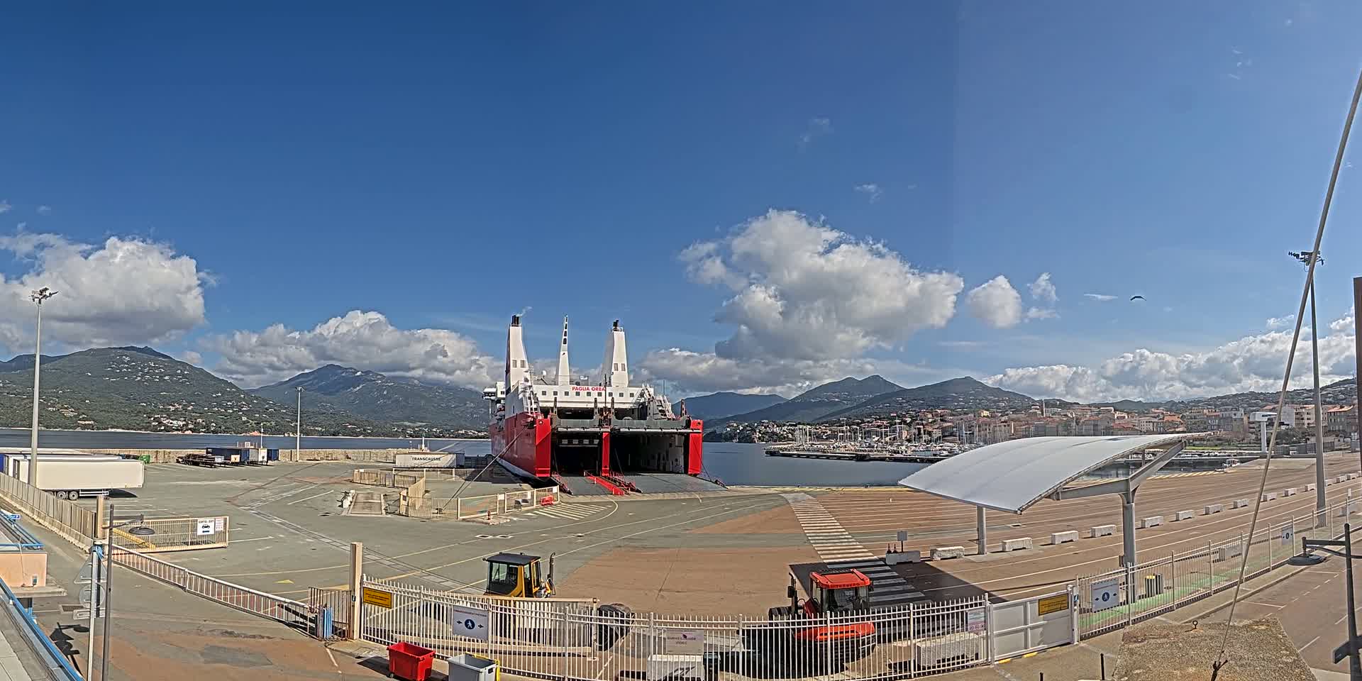 A large red and white ferry is docked at a port, under a partly cloudy sky, with mountains in the background.