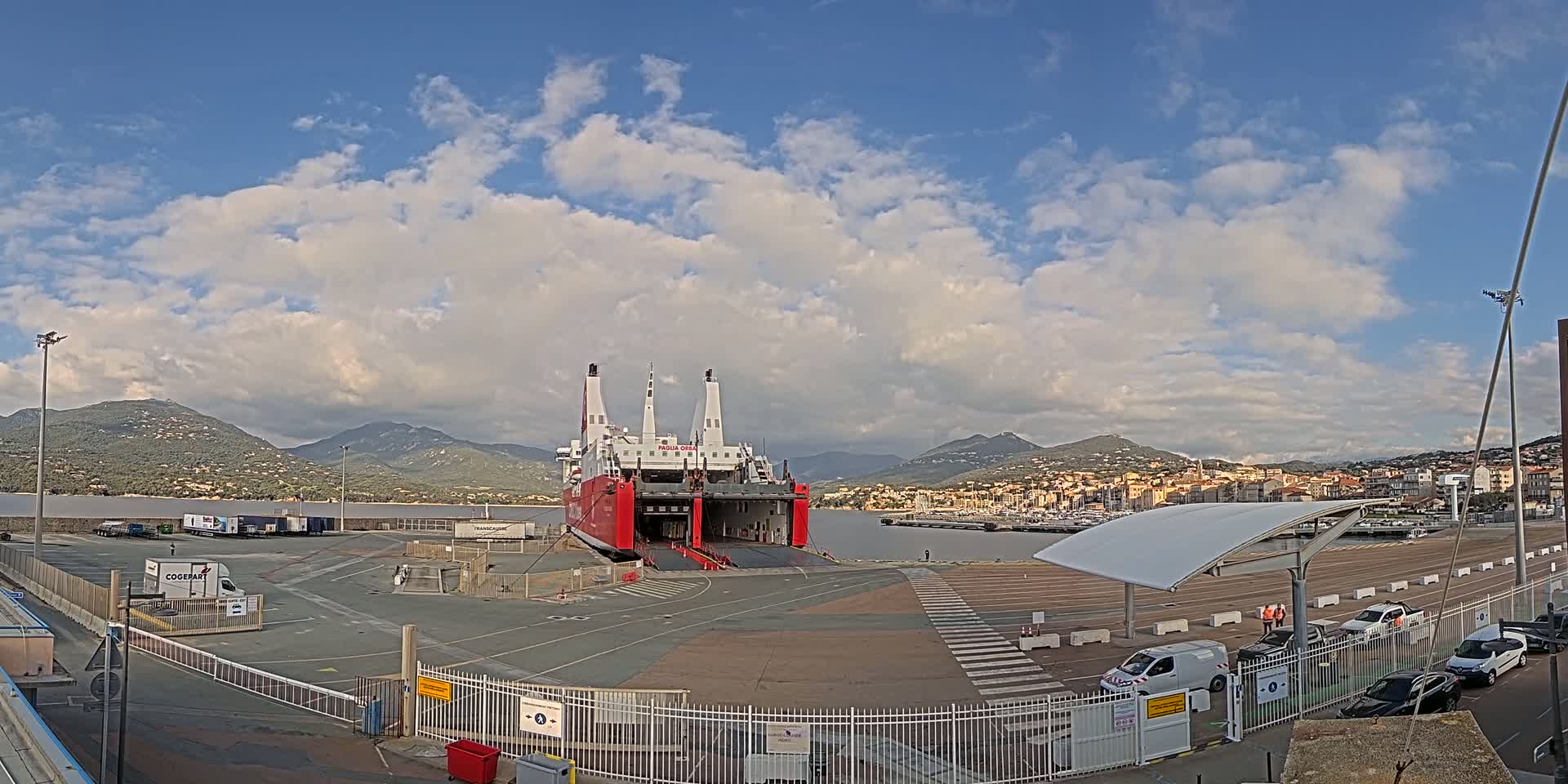 A large red and white ferry is docked at a port under a partly cloudy sky, with mountains and a town visible in the background.
