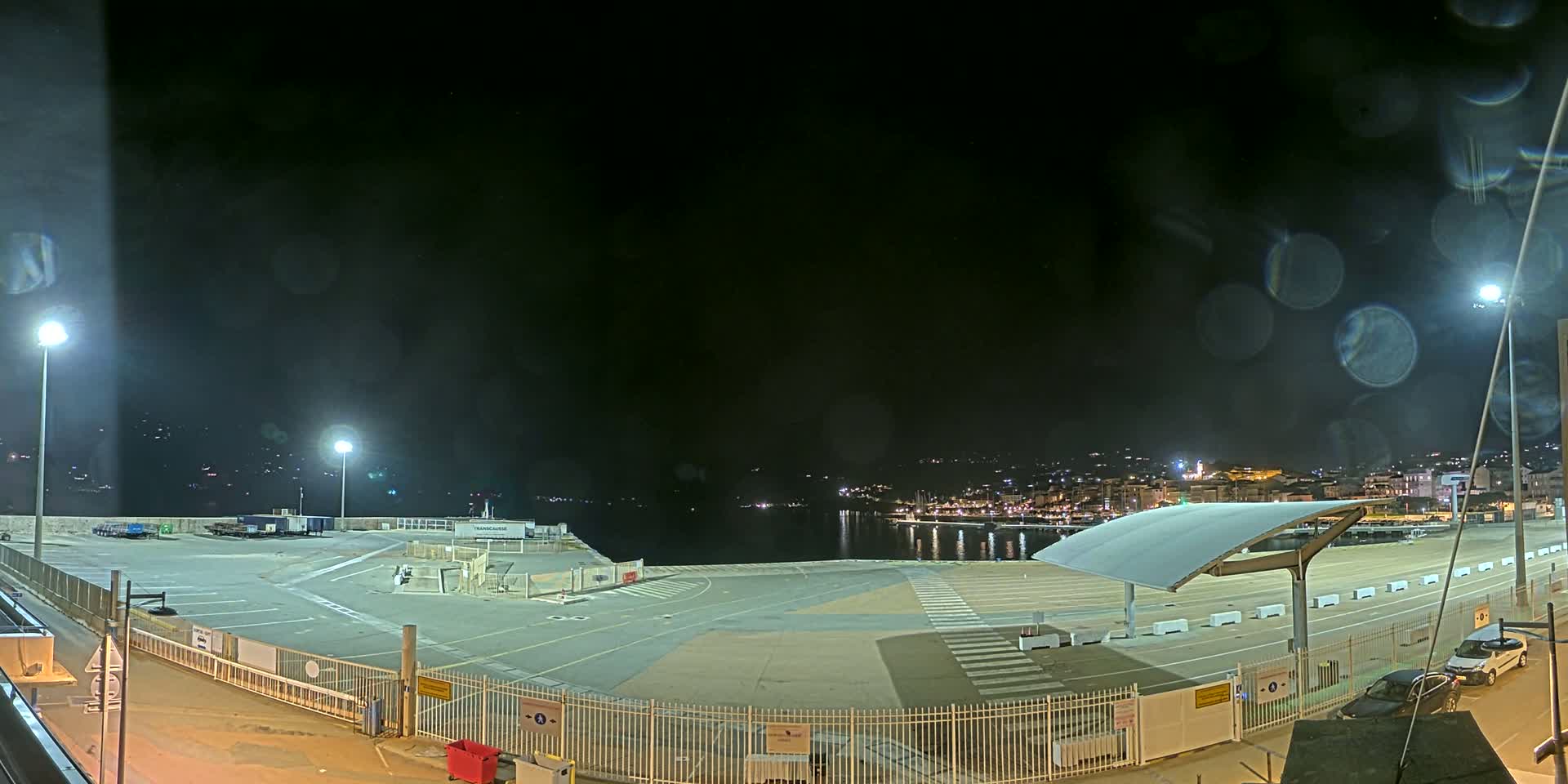 An empty, illuminated nighttime port area with a few parked cars, viewed through a rain-splattered window, overlooks a dark, calm body of water and a distant, softly lit town.
