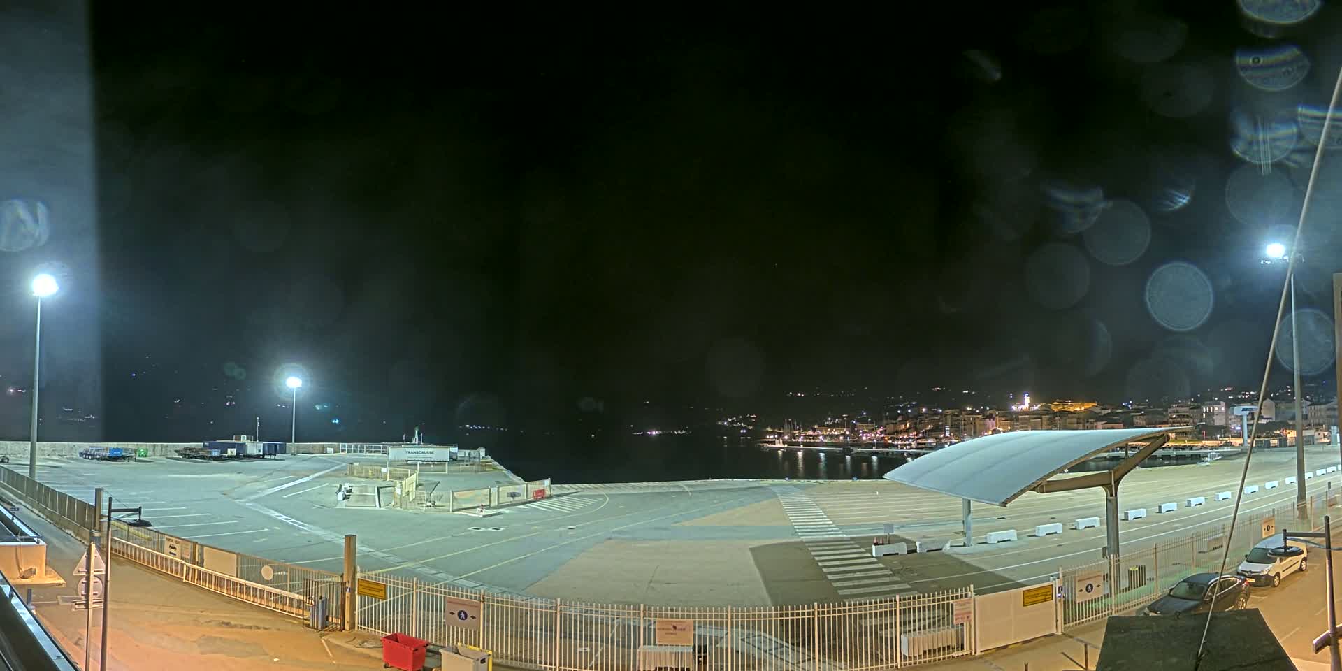 A nighttime view of an empty port area with a few parked cars, illuminated by lights, under a dark, possibly rainy sky.