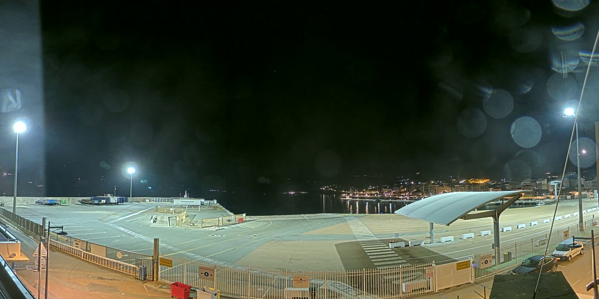 An empty, nighttime port area with a few parked cars, illuminated by several lights, overlooks a calm body of water with a town visible in the distance under a dark, slightly misty sky.