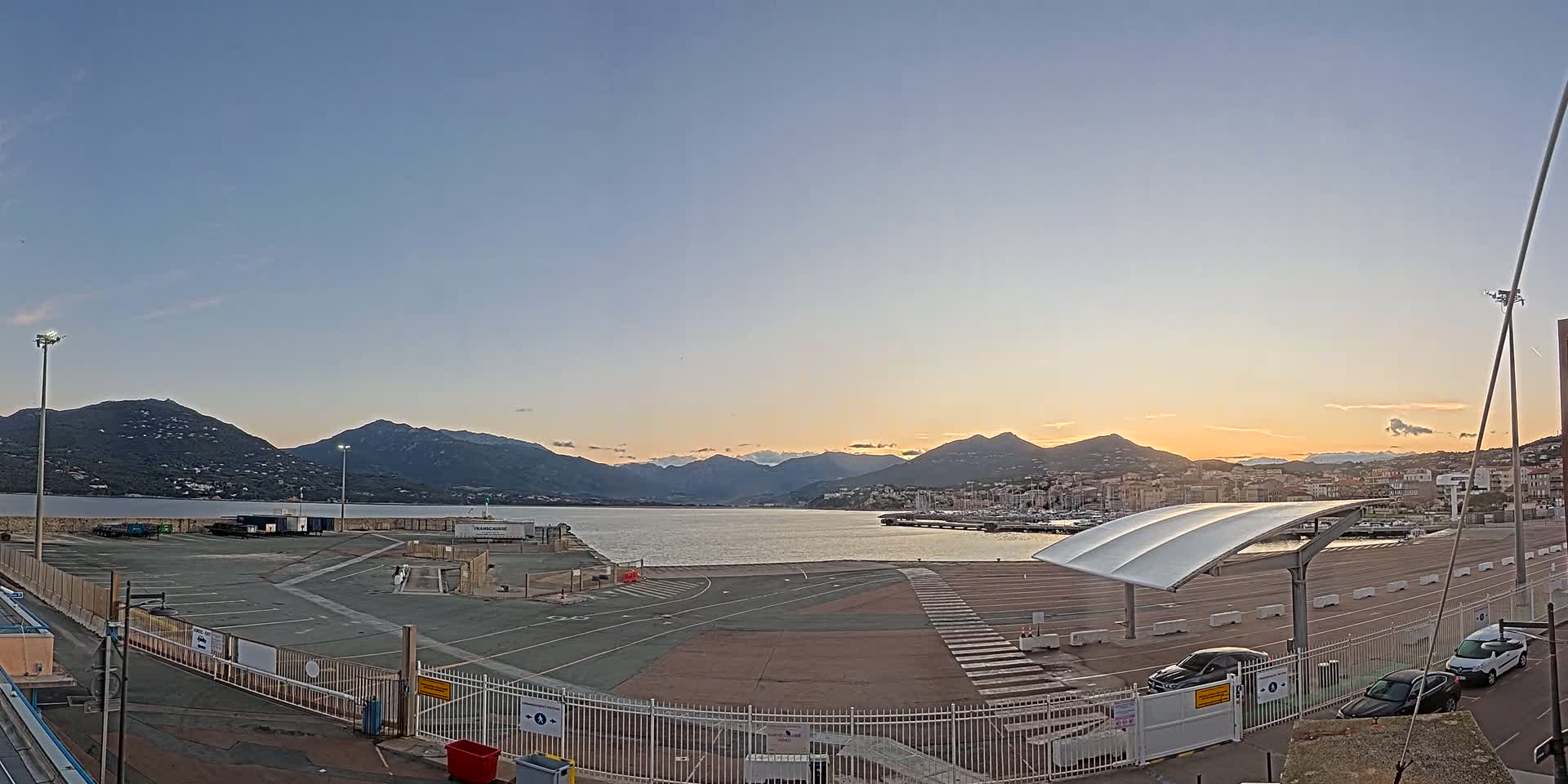 An empty port area with a few cars, under a clear sunset sky, overlooks a calm body of water and mountains in the distance.