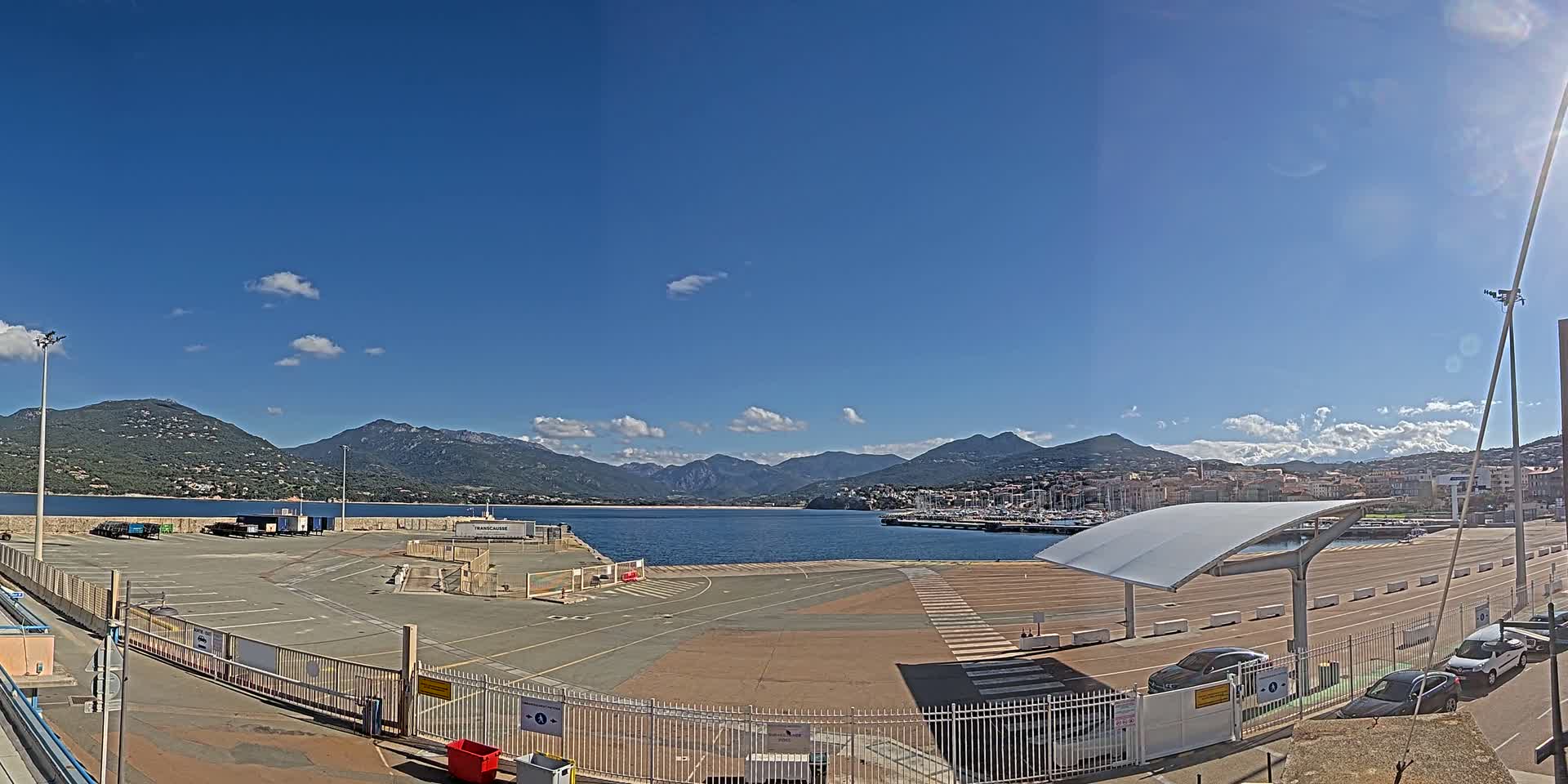 A mostly sunny day shows a port with an empty parking lot, a covered waiting area, and a distant view of mountains and a town across the water.