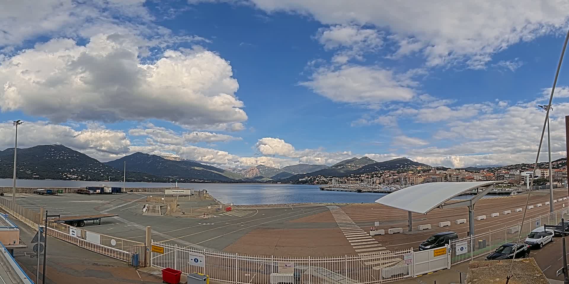 A partly cloudy day reveals a harbor's empty parking lot and a town nestled against mountains in the distance.
