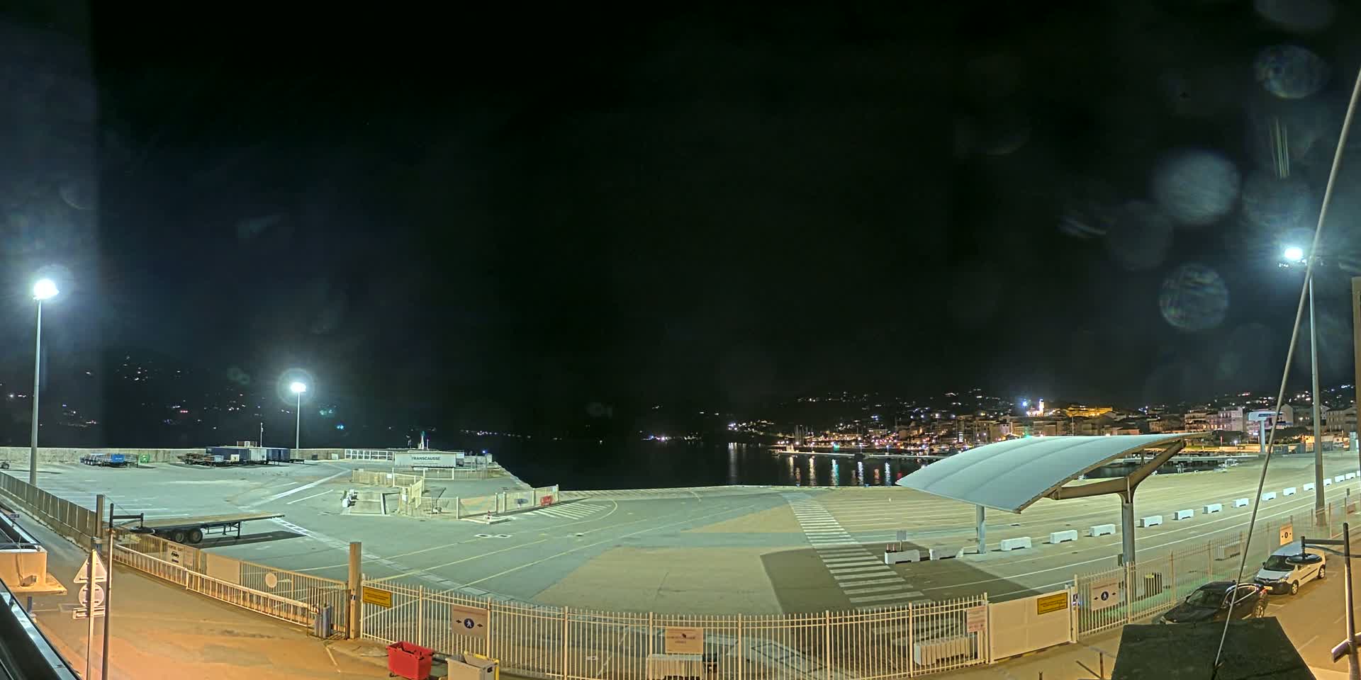 A nighttime view of an empty port area with a few parked vehicles, illuminated by several streetlights, overlooking a dark body of water and a city skyline in the distance.