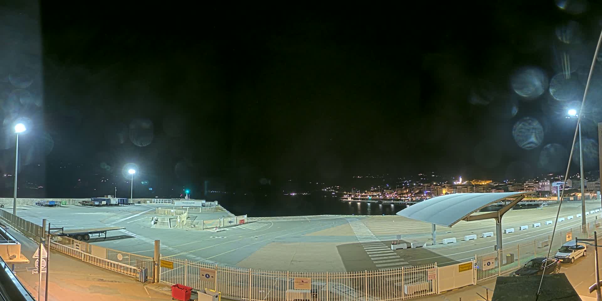 An empty, brightly lit port area at night, with rain obscuring the view, overlooks a dimly lit town across the water.