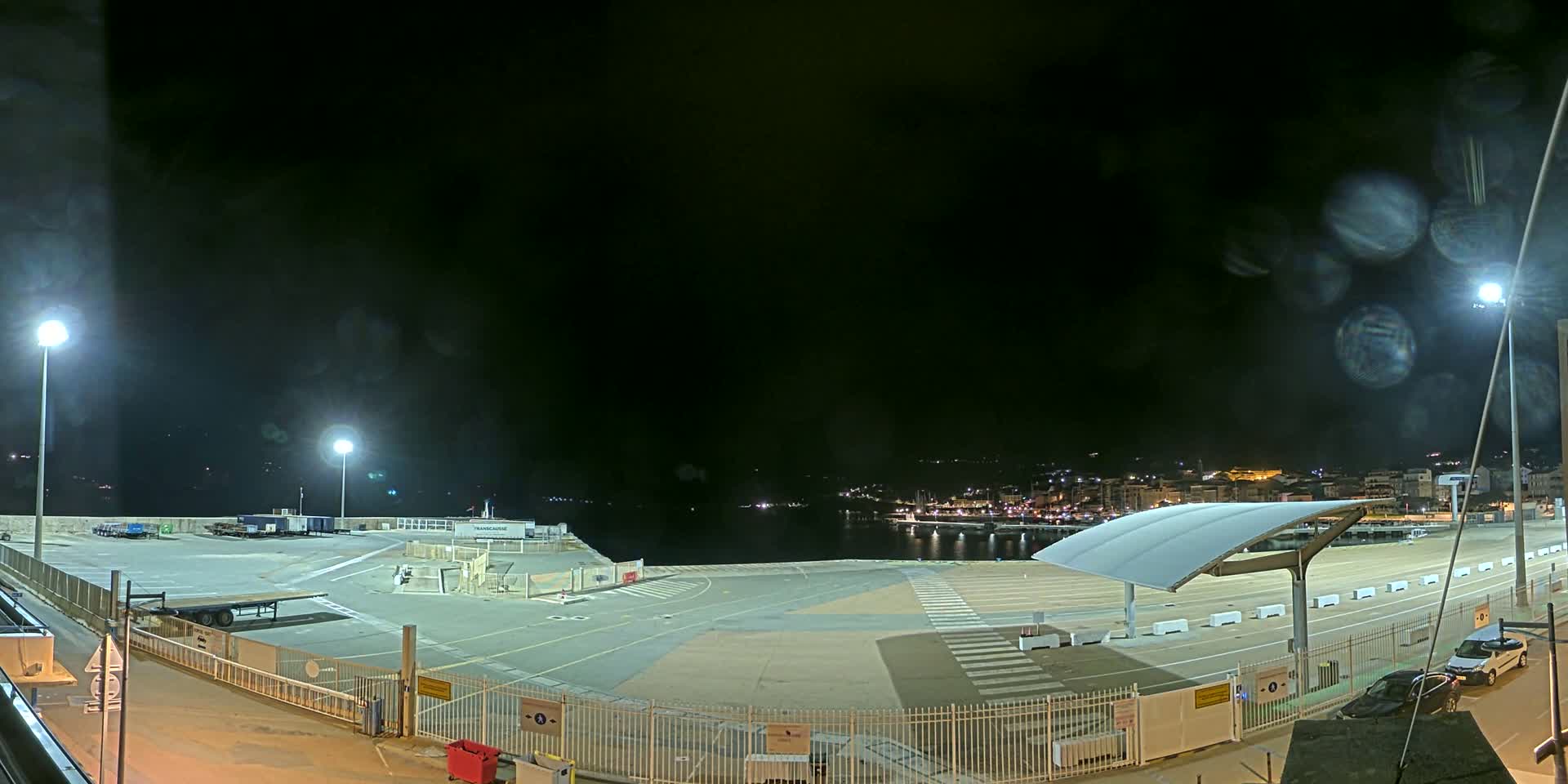 A nighttime view of an empty port area with a few parked vehicles, lit by several bright lamps, under a dark, possibly slightly misty sky.