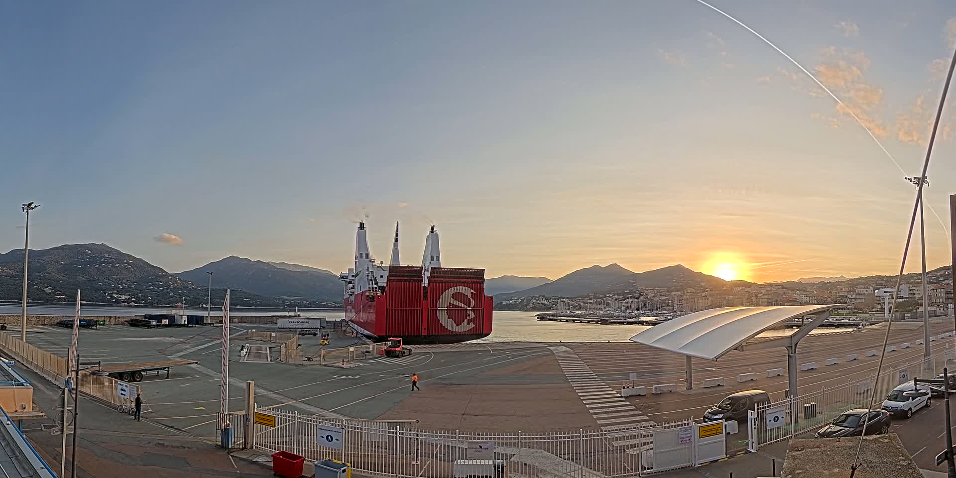 A large red cargo ship is docked at a port during a clear sunset, with mountains visible in the background.
