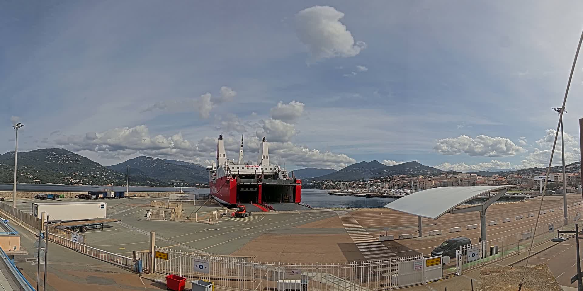A large red and white ferry is docked at a port, under a partly cloudy sky, with mountains in the background.