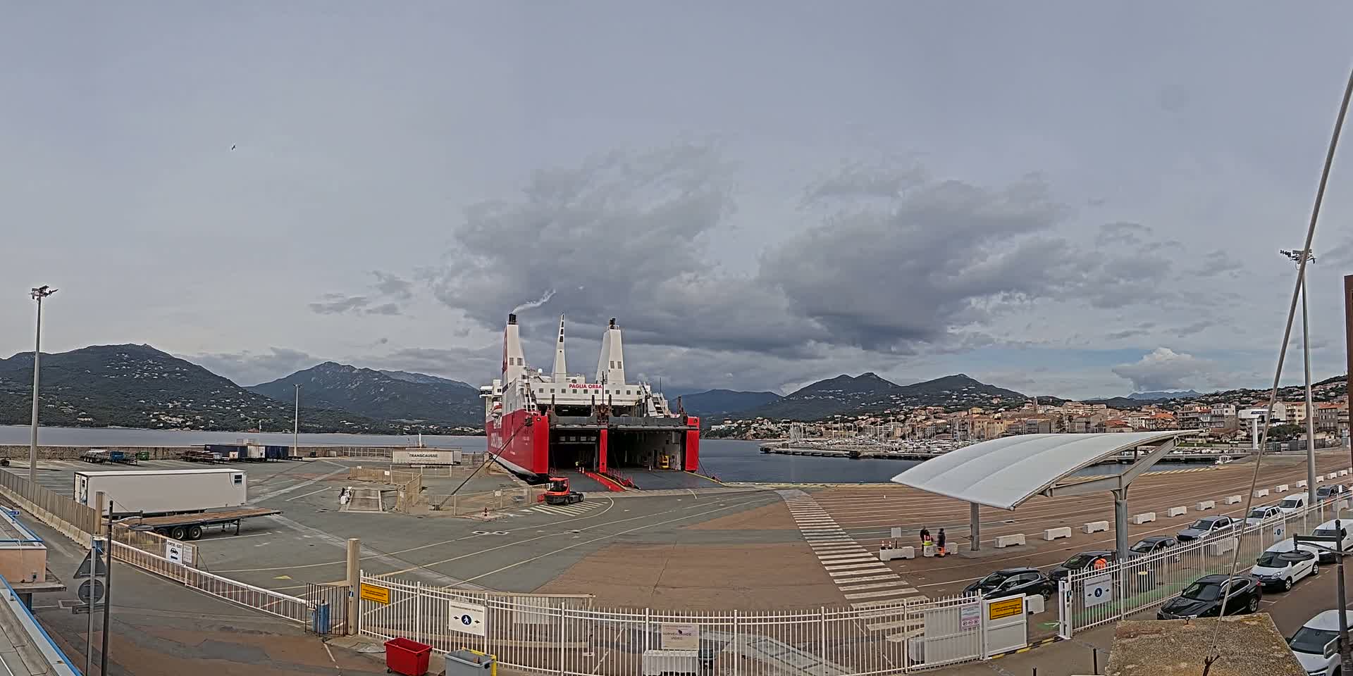 A large red and white ferry is docked at a port, under a cloudy sky, with mountains in the background.
