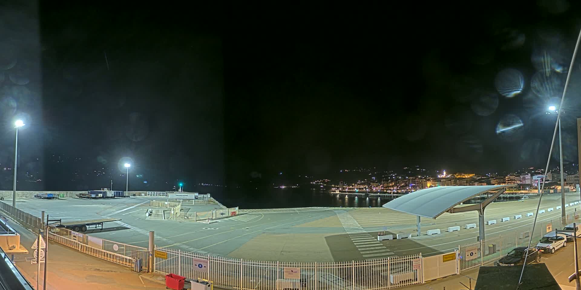 A nighttime view of a mostly empty port area with a town's lights visible across the water under a dark, possibly slightly hazy sky.