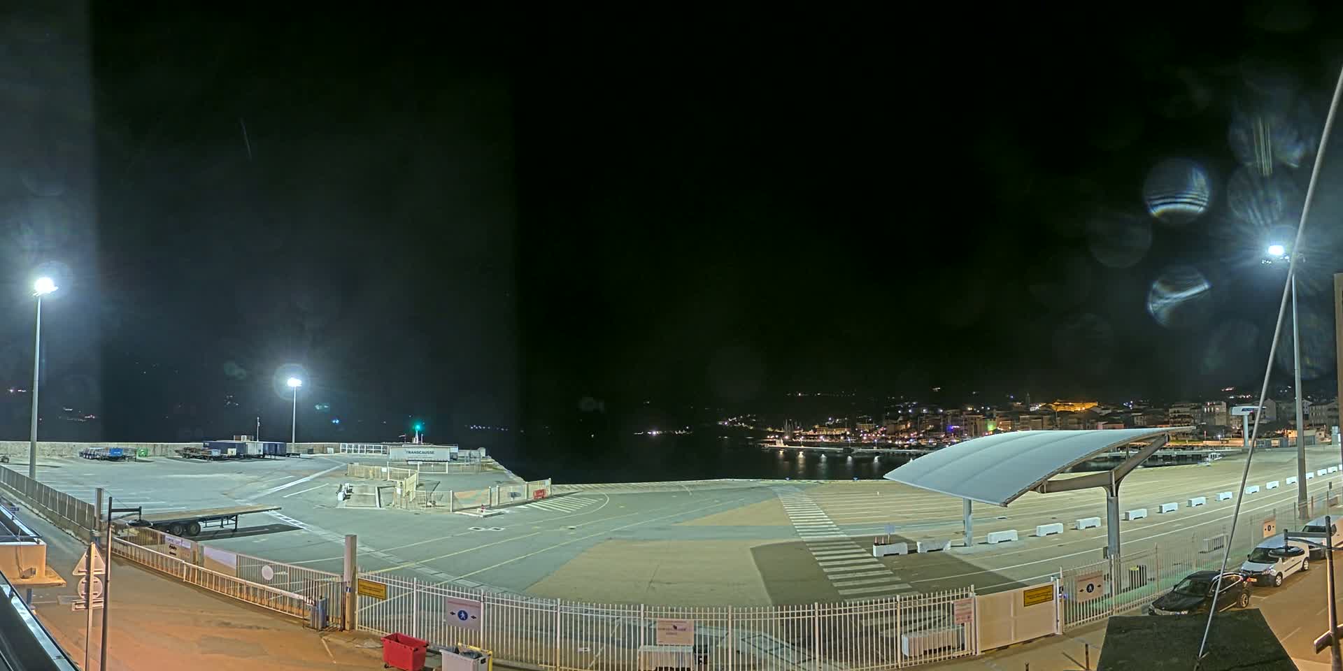 A nighttime view of an empty port area with a few vehicles parked near a fence, overlooking a calm body of water with a town illuminated in the distance.