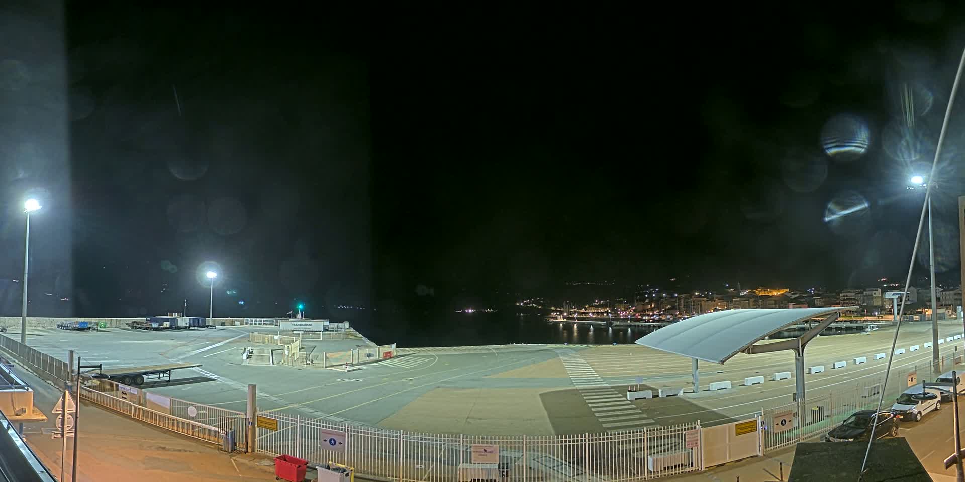 A nighttime view of an empty port area with a few parked cars, illuminated by several lights, under a slightly hazy sky.