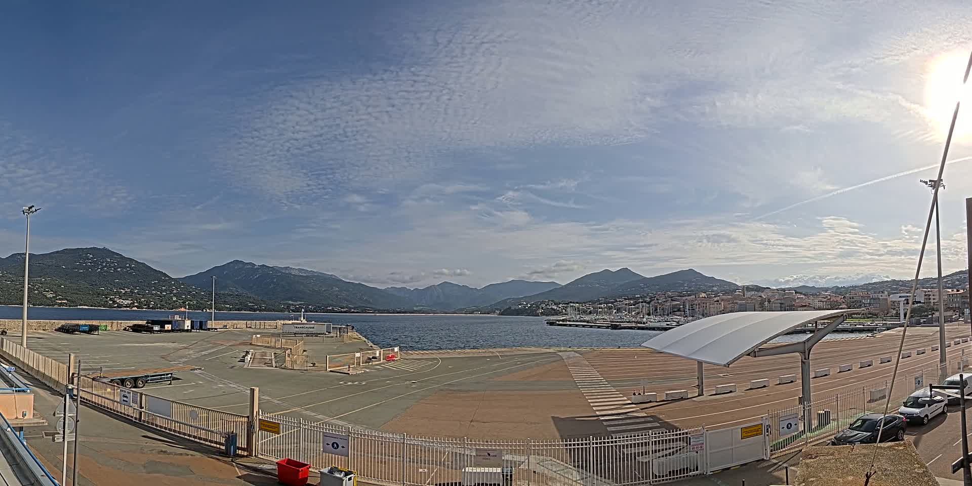 A mostly sunny day shows a port area with a large paved lot,  a few vehicles, and mountains in the background across a calm body of water.