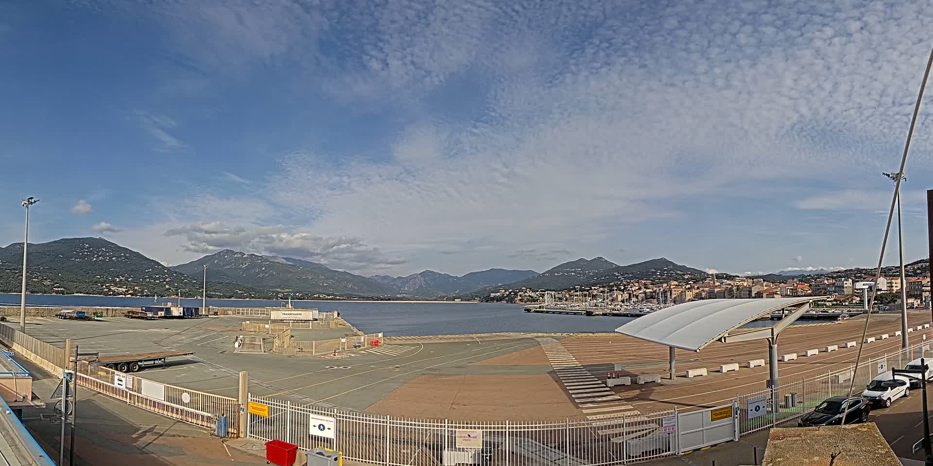 A mostly sunny day shows a port area with trucks, cars, and a covered waiting area, overlooking a calm body of water and mountains in the distance.