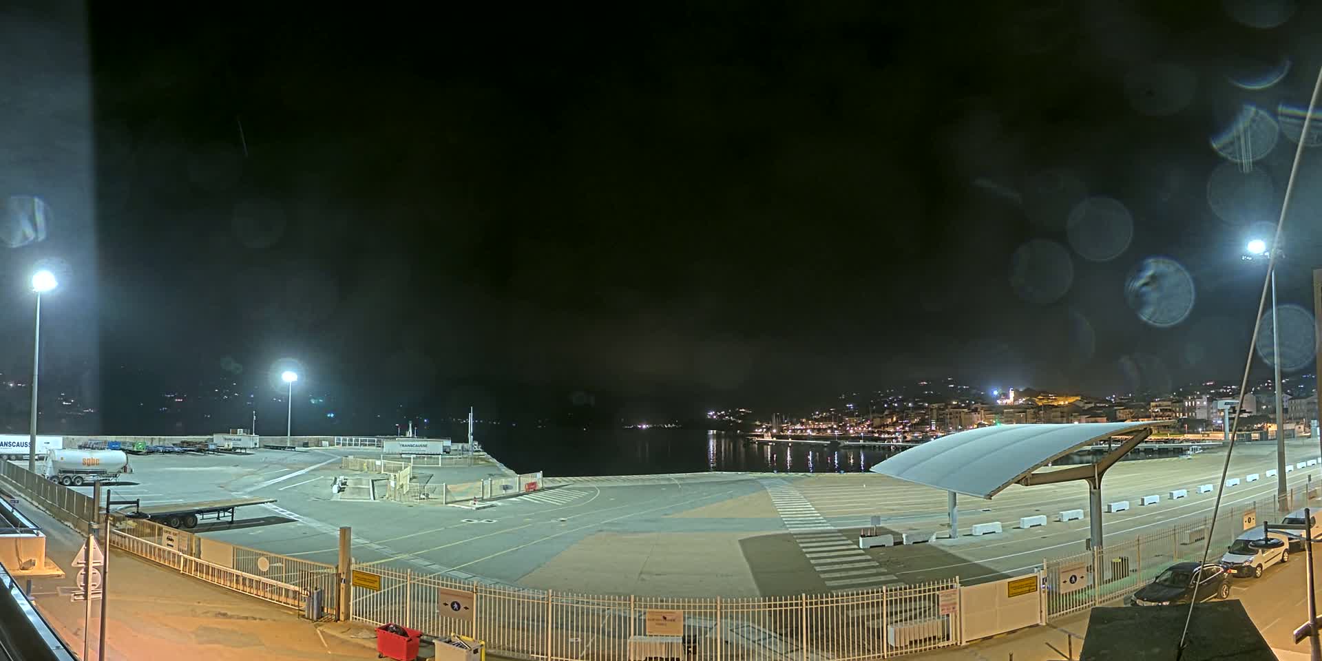 A nighttime view of a port area with a mostly empty paved lot,  a few parked cars, and a town visible across the water, under a dark, rainy sky.