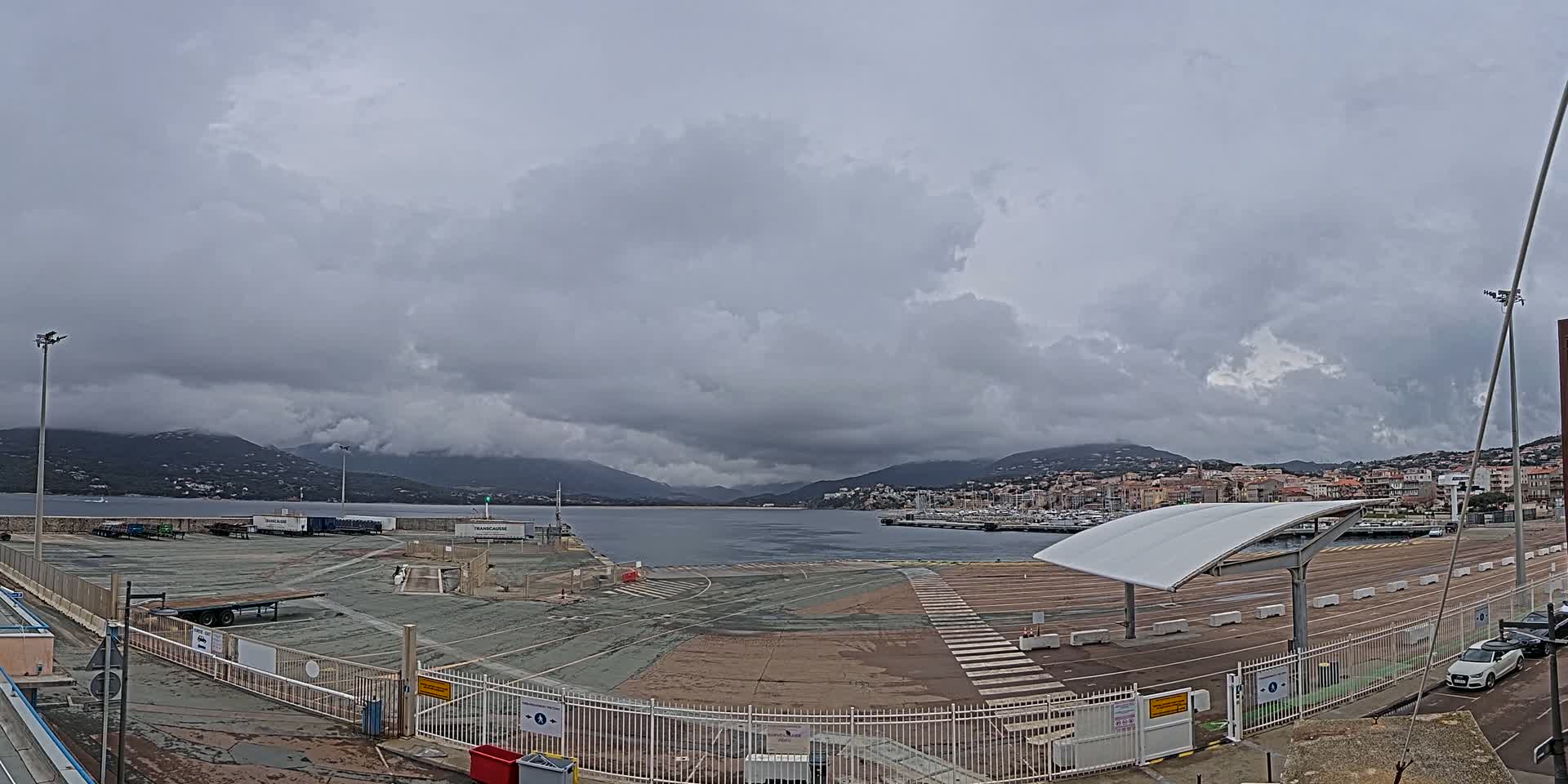 An overcast day reveals a port area with a large empty paved lot, a marina with boats, a town in the distance, and mountains beyond under a cloudy sky.