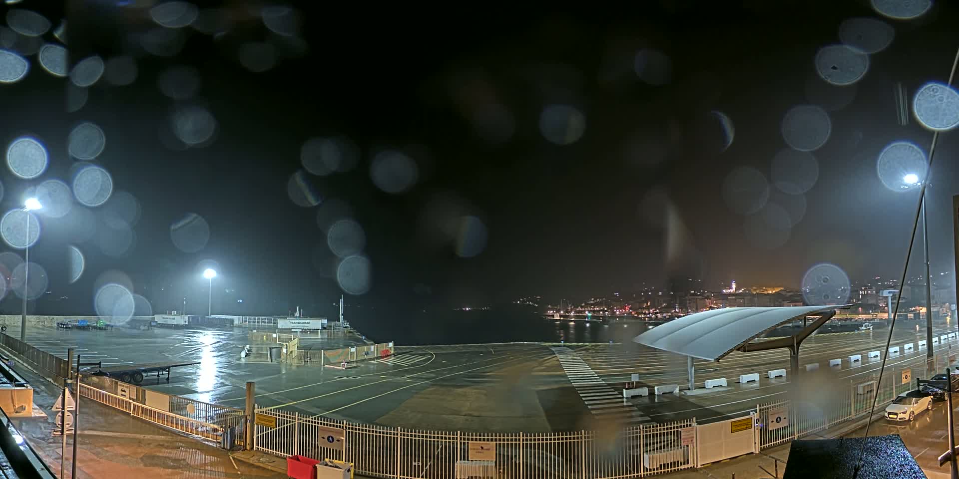A rain-soaked, nighttime view of a port area with empty parking lots, illuminated by several bright lights, and a city skyline in the distance.