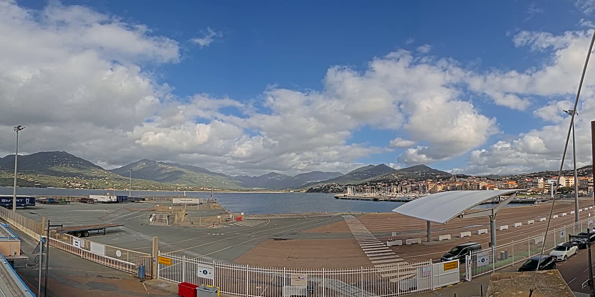 A mostly sunny day with partly cloudy skies over a port area with a large paved lot, a marina with boats, and mountains in the background.