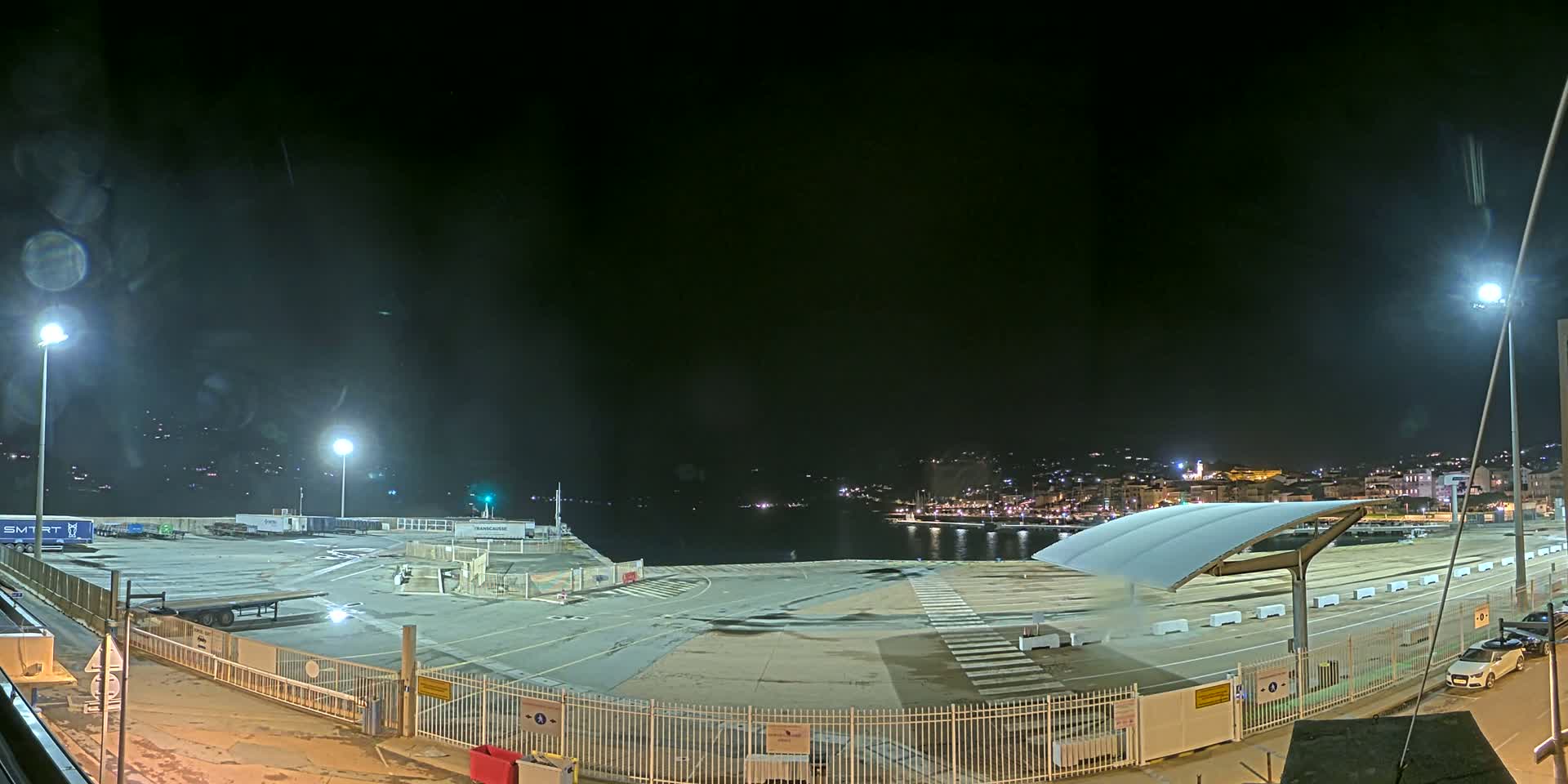 A nighttime view of a mostly empty port area with a few vehicles and containers, illuminated by artificial lights, overlooking a dark body of water and a city skyline in the distance.